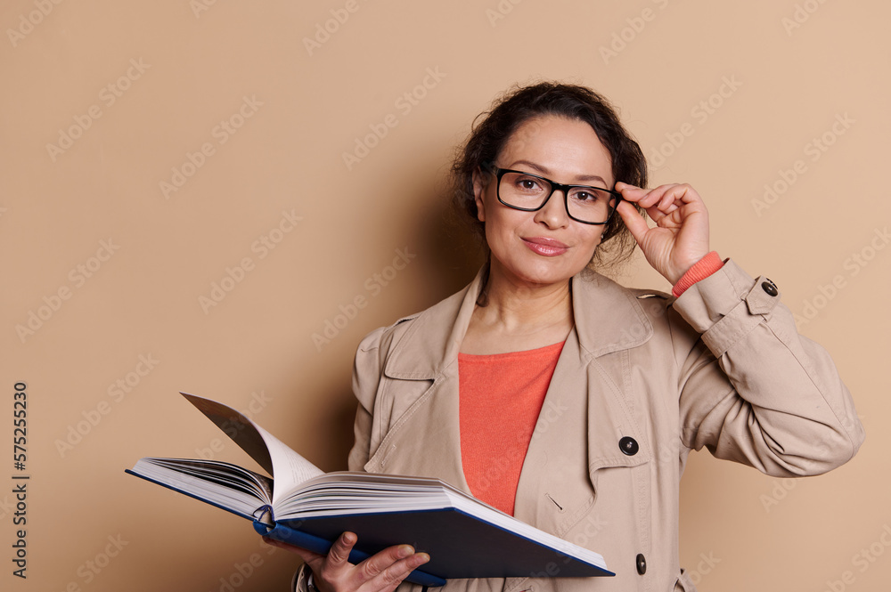Multi-ethnic beautiful middle-aged woman, a teacher, professor, educator, holding a hardcover book with fairy tails, and smiling looking at camera through eyeglasses, isolated beige color background Multi-ethnic beautiful middle-aged woman, a teacher, professor, educator, holding a hardcover book with fairy tails, and smiling looking at camera through eyeglasses, isolated beige color background