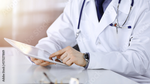 Unknown male doctor sitting and working with tablet computer iin a darkened clinic, glare of light on the background, close-up of hands