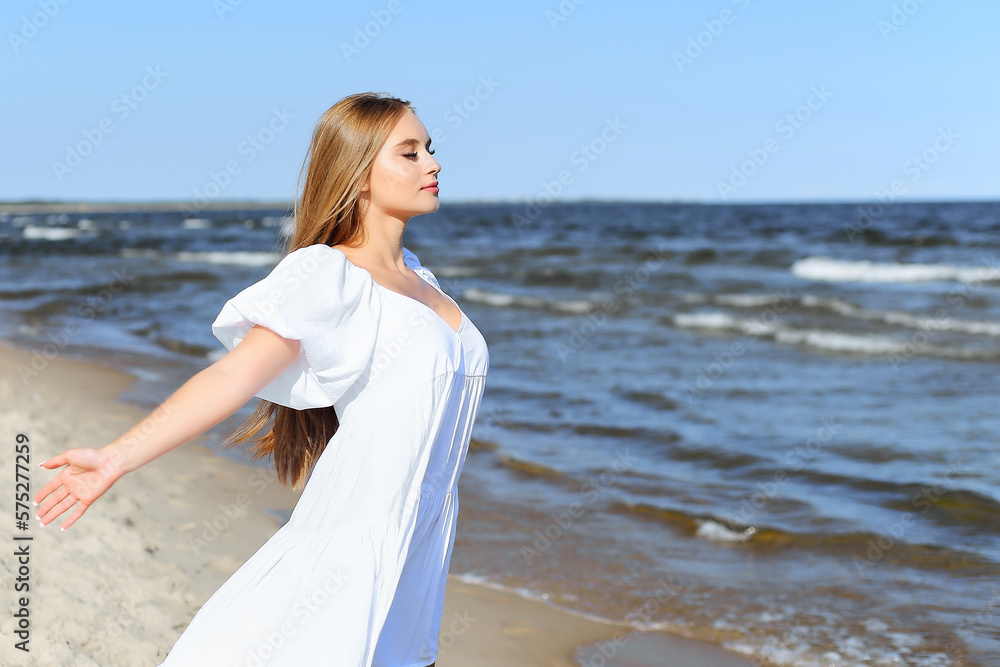 Happy smiling beautiful woman is on the ocean beach in a white summer dress, open arms