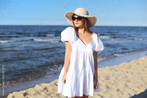 Happy smiling blonde woman is posing on the ocean beach with sunglasses and a hat