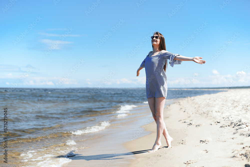 Happy smiling woman in free bliss on ocean beach standing with open hands. Portrait of a brunette female model in summer dress enjoying nature during travel holidays vacation outdoors, overall plan