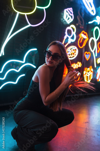 Beautiful fashionable woman in an elegant black dress with sunglasses is sitting and fixing her hair on a dark background with multicolored neon signs lights