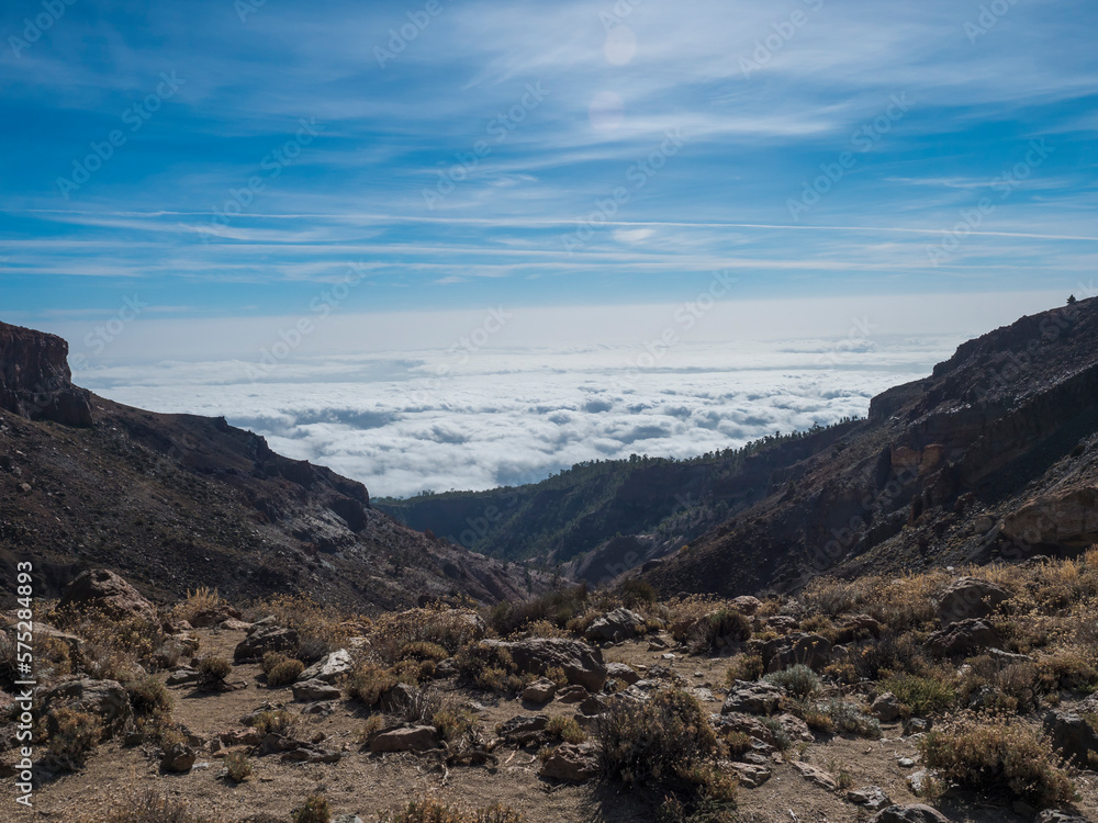 View from mountain pass of Alto de Guajara volcano with with lava rock boulders and dry vegetation, above the white clouds, blue sky background. El Teide National Park, Tenerife, Canary islands, Spain