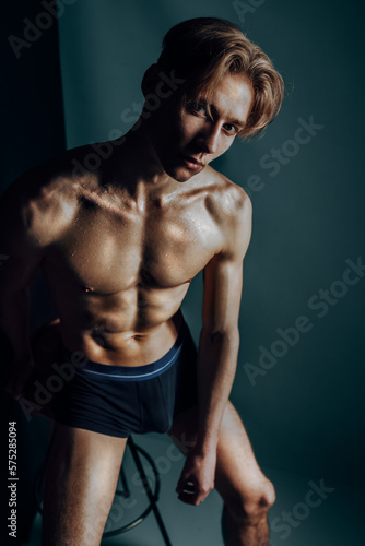hot male model sit nude and erotic in dark blue studio on stool express his masculine body shaped by fitness and aesthetics as symbol for traditional gender expression while being macho and sex symbol