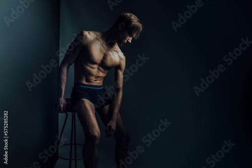 hot male model sit nude and erotic in dark blue studio express his masculine body shaped by fitness and aesthetics as symbol for traditional gender expression while being macho and sex symbol