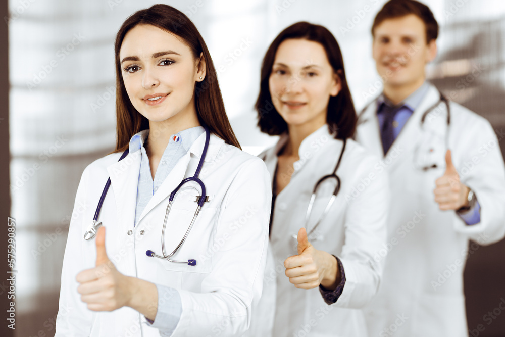 Group of young cheerful doctors is standing as a team with thumbs up in a hospital office and is ready to help patients. Medical help, insurance in health care Group of young cheerful doctors is standing as a team with thumbs up in a hospital office and is ready to help patients. Medical help, insurance in health care