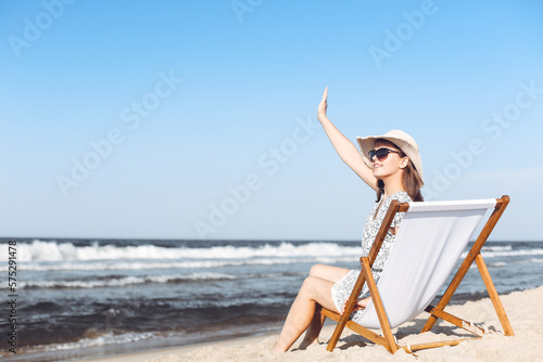 Happy brunette woman sitting on a wooden deck chair at the ocean beach while waving and greeting somebody with her hand