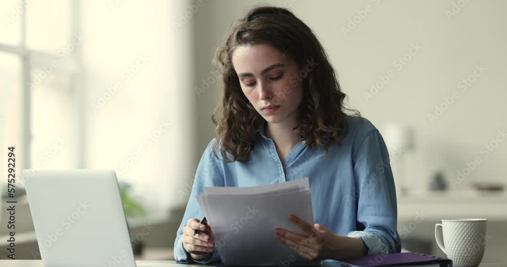 Young 20s thoughtful woman working with papers or studying, reading essay, learning, check written information, prepare assignment, making test, looks concentrated sits at desk with laptop. Paperwork Young 20s thoughtful woman working with papers or studying, reading essay, learning, check written information, prepare assignment, making test, looks concentrated sits at desk with laptop. Paperwork