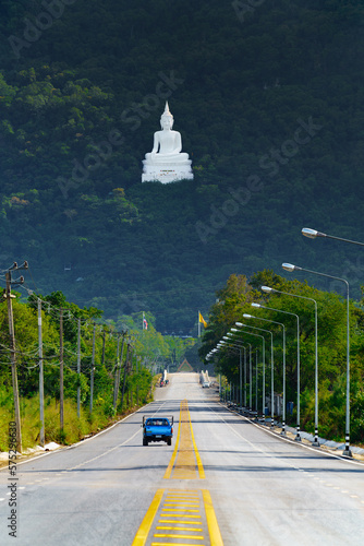 Buddha Sakolsima Mongkhon at Temple  Wat Thep Phithak Punnaram. Large white Buddha sits high in the Forest Hill viewed from a distance down on the road with a blue pick up truck driving.