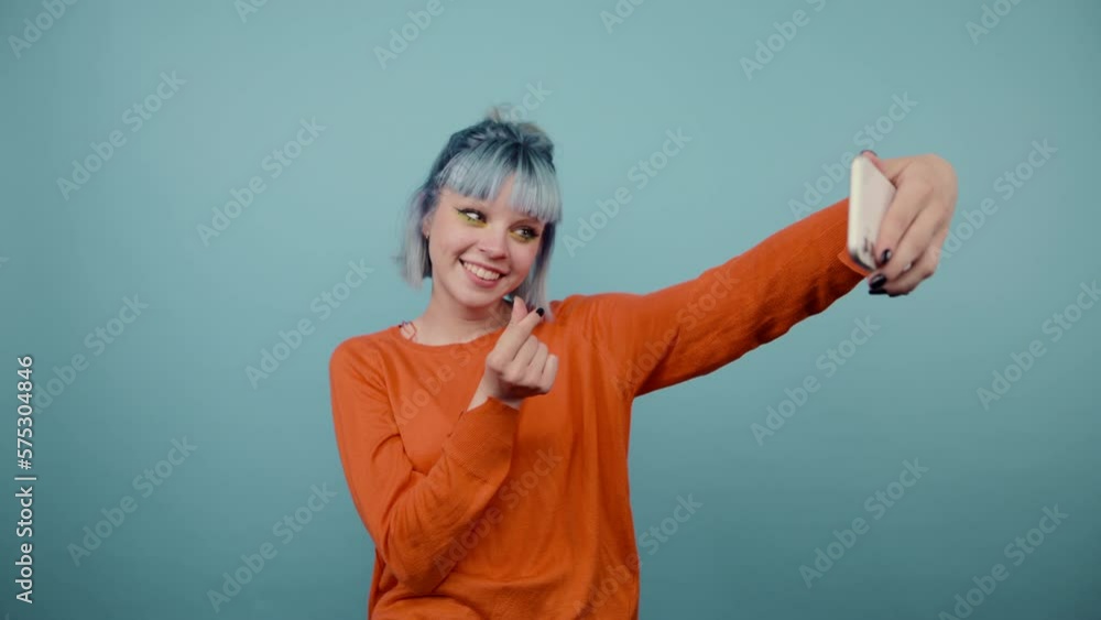 Positive teenage girl with colored hair taking selfie on blue background and showing heart gesture. Positive teenage girl with colored hair taking selfie on blue background and showing heart gesture.