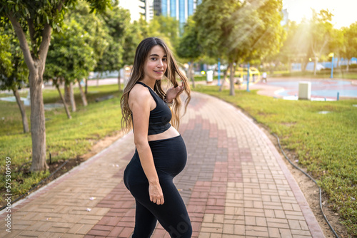 Portrait of nine month pregnant woman in sportswear holding her hair and standing on park sidewalk.