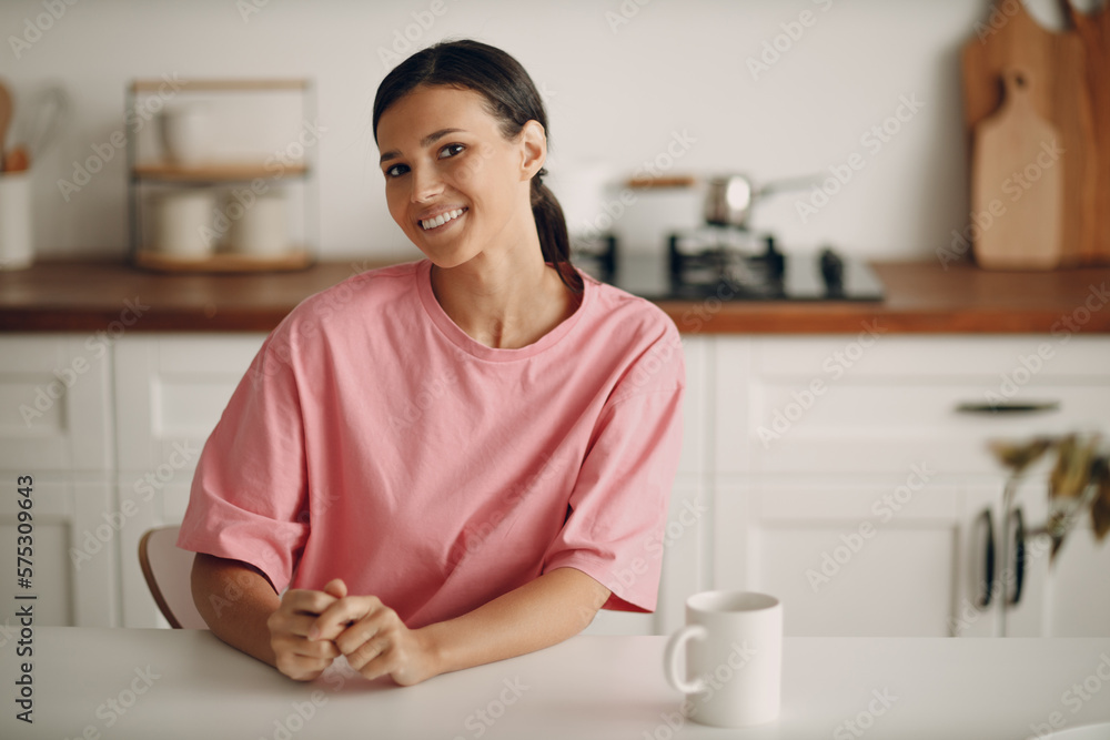 Cute young brunette woman in casual clothes looks straight into the camera. Portrait of smiling happy girl sitting at home in the kitchen at the dining table. Diet, fasting. Cute young brunette woman in casual clothes looks straight into the camera. Portrait of smiling happy girl sitting at home in the kitchen at the dining table. Diet, fasting.