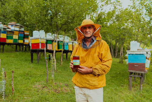 The beekeeper holding a jar of honey in his hand while standing in a meadow surrounded by a box and a honey farm