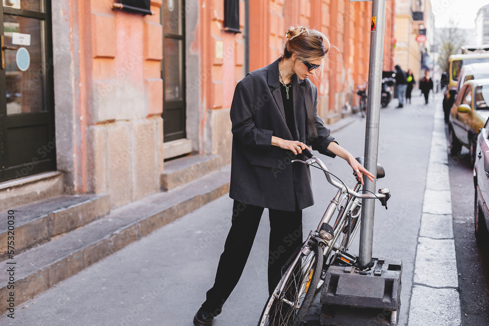 Attractive business woman come to sits on bike. Girl wear grey jacket or blazer, black pants, eyeglasses. Modern business woman ride to work on vehicle. Way to office. Riding bicycle to work. Attractive business woman come to sits on bike. Girl wear grey jacket or blazer, black pants, eyeglasses. Modern business woman ride to work on vehicle. Way to office. Riding bicycle to work.