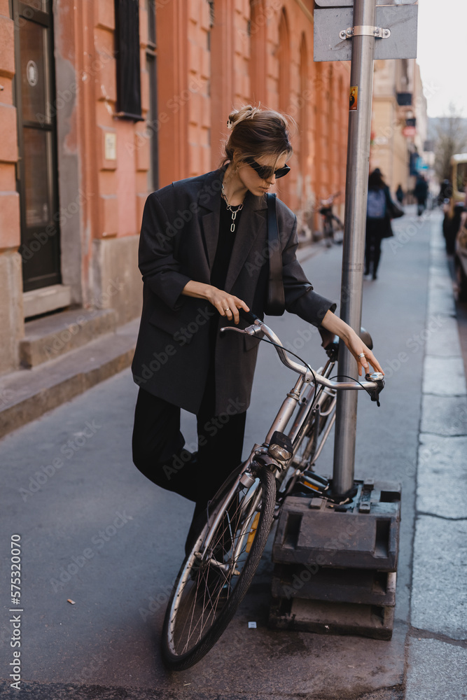 Attractive business woman come to sits on bike. Girl wear grey jacket or blazer, black pants, eyeglasses. Modern business woman ride to work on vehicle. Way to office. Riding bicycle to work.