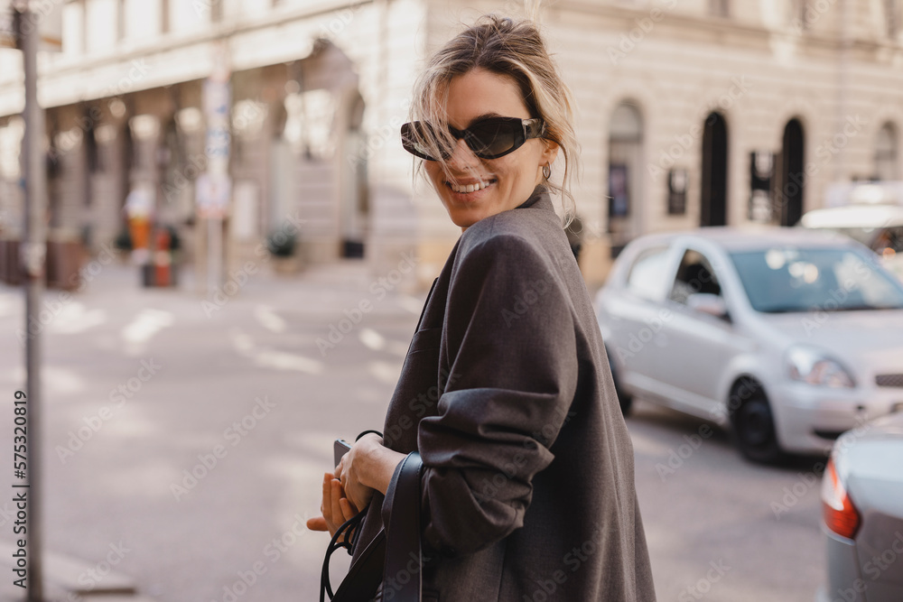 Young beautiful smiling hipster woman in trendy outfit. Sexy carefree woman posing on the street. Cheerful and happy in black sunglasses and grey suit turn around. Lifestyle, female beauty concept.