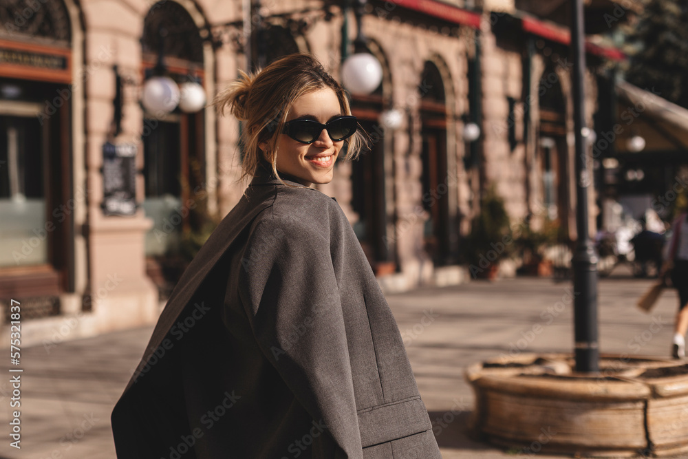 Young beautiful smiling hipster woman in trendy outfit. Sexy carefree woman posing on the street. Cheerful and happy in black sunglasses and grey suit turn around. Lifestyle, female beauty concept. Young beautiful smiling hipster woman in trendy outfit. Sexy carefree woman posing on the street. Cheerful and happy in black sunglasses and grey suit turn around. Lifestyle, female beauty concept.