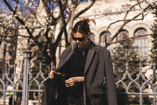 Elegant young woman looking in her black leather bag her phone or purse. Business style woman wear grey blazer, black eyeglasses and bag on the street. Street style, fashion outfit.