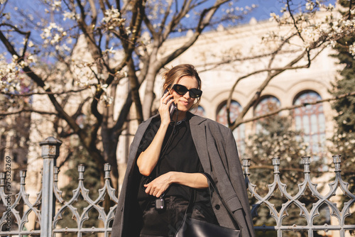 Beautiful young woman wearing suit, walking outdoors, talking on mobile phone. Beautiful Woman talking on phone. Portrait of stylish smiling business woman calling on mobile phone under blooming tree.