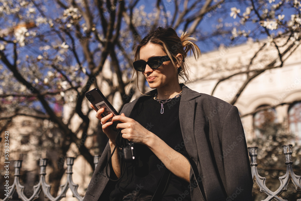 Happy woman walking using smart phone in a city street. Business woman with sunglasses uses mobile phone outdoors under blooming tree, girl typing message, chatting, scrolling web page, read good news