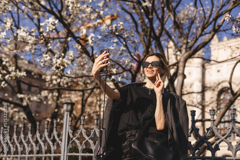Beautiful smiling blonde model in summer or spring formal clothes. Carefree female posing in sunglasses. Taking selfie self portrait photos on smartphone, shows peace sign, kiss. Focus on peace sign. Beautiful smiling blonde model in summer or spring formal clothes. Carefree female posing in sunglasses. Taking selfie self portrait photos on smartphone, shows peace sign, kiss. Focus on peace sign.