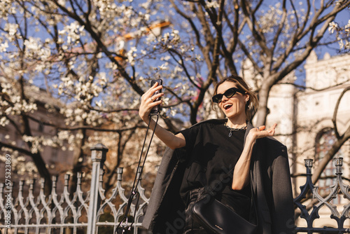 Happy young woman photographing herself using her mobile phone. Caucasian female talking selfie with her smart phone on blooming tree background. Girl posing in sunglasses, suit, t-shirt, pants, bag.