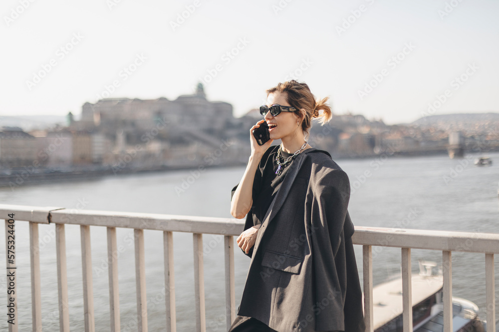 Woman talking on the phone walking on the bridge under the river. Beautiful young woman with blond hair smiling and having a conversation over the phone in the city. Business woman on smartphone.