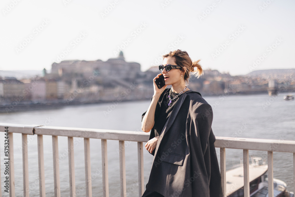 Woman talking on the phone walking on the bridge under the river. Beautiful young woman with blond hair smiling and having a conversation over the phone in the city. Business woman on smartphone.