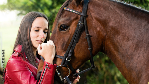 Selective focus of pensive woman stroking her brown horse in nature. Woman with brown horse having special connection with animal. Thoughtful young woman with hands on horse's head with trees behind.