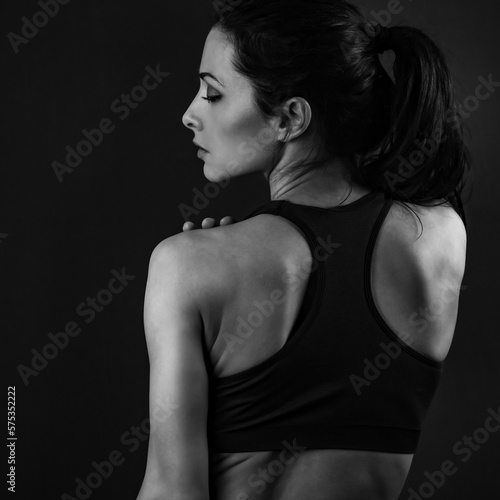 Serious female sporty muscular with ponytail doing stretching workout the shoulders, blades and arms in sport bra, standing on dark shadow grey background with empty copy space. Back view. Lifestyle