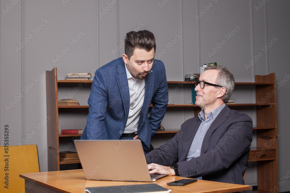 Mature businessman in blue suit and his younger colleague are thinking about business plans and discussing them in office Mature businessman in blue suit and his younger colleague are thinking about business plans and discussing them in office