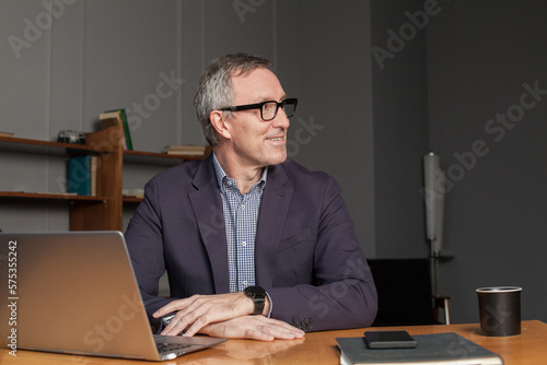 Mature happy businessman or entrepreneur in eye glasses and suit is smiling sitting by the laptop in office
