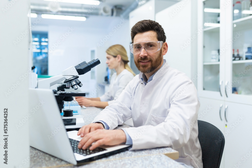 Portrait of mature successful scientist lab technician at workplace in laboratory with microscope, senior man with beard and goggles smiling and looking into camera, inventor with laptop .