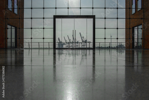 Blick durch ein großes modernes Glasfenster, mittig eine große Glastür, links und rechts rotbraune Gebäudeteile, im Hintergrund die Silhouetten von Hafenkränen und Industriegebäuden zu sehen, horizont photo