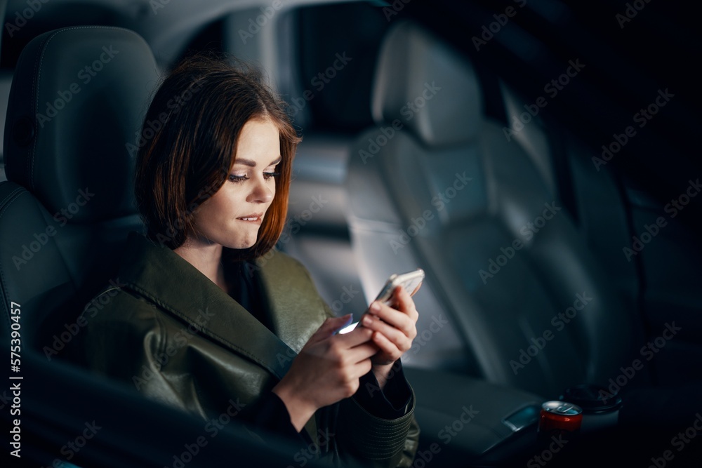 horizontal portrait of a stylish, luxurious woman in a green leather coat, sitting in a black car at night in the passenger seat, playfully biting her lower lip, holding her phone during the trip