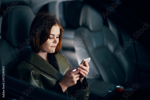 horizontal portrait of a stylish, luxurious woman in a green leather coat, sitting in a black car at night in the passenger seat, playfully biting her lower lip, holding her phone during the trip © SHOTPRIME STUDIO