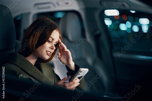 a close horizontal portrait of a stylish, luxurious woman in a leather coat sitting in a black car at night in the passenger seat, thoughtfully looking at her smartphone with her hand near her face © SHOTPRIME STUDIO