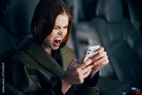 a close horizontal portrait of a stylish, luxurious woman in a leather coat sitting in a black car at night in the passenger seat, emotionally looking into her smartphone with her mouth wide open © SHOTPRIME STUDIO