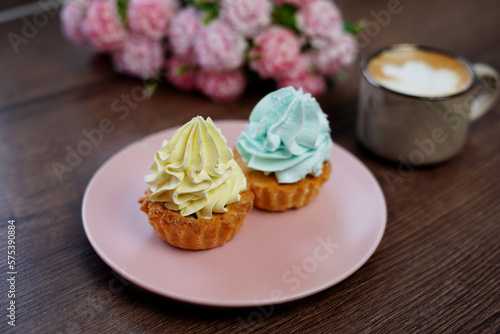 A Pink Plate with birthday Capcakes sits on a wooden table. A beige cup of cappuccino sits on the table. Near a plate of cakes lie beautiful pink and white flowers.