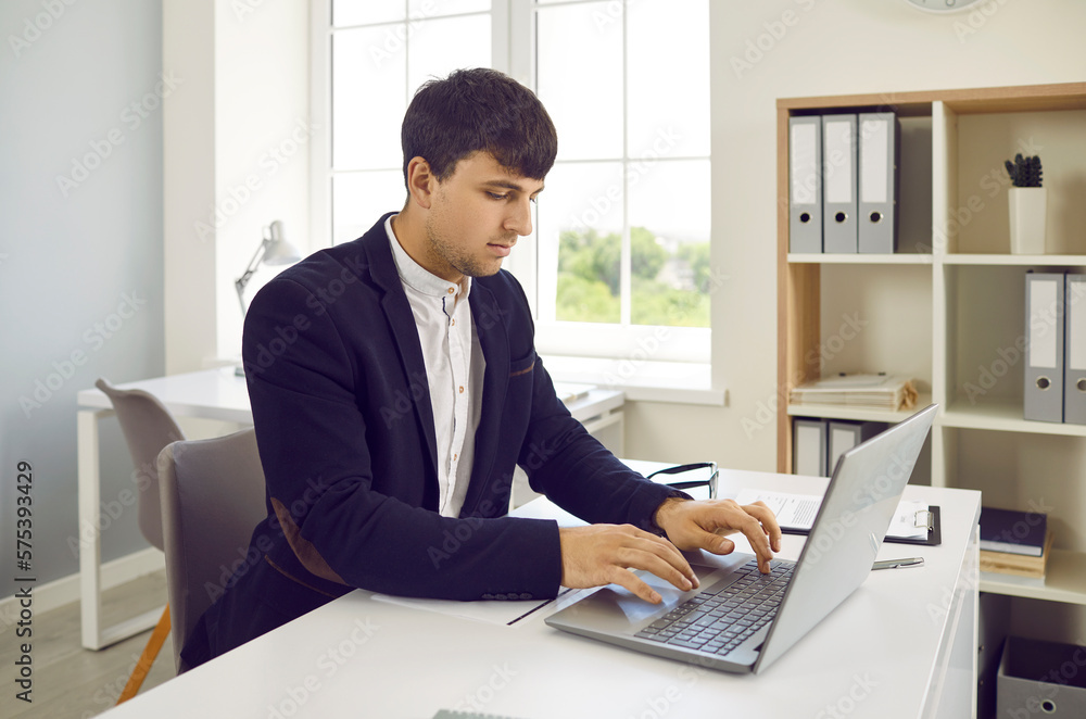 Young businessman working in office. Young man sitting at his office desk, typing on laptop computer, searching for information, developing online his business and working with digital documents Young businessman working in office. Young man sitting at his office desk, typing on laptop computer, searching for information, developing online his business and working with digital documents
