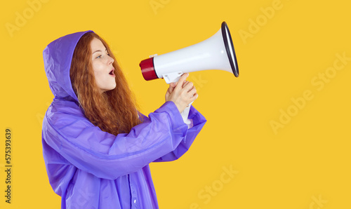 Canvas Print Funny red-haired beautiful young woman, dressed in waterproof, sings with her mouth open in megaphone, holding it with both hands