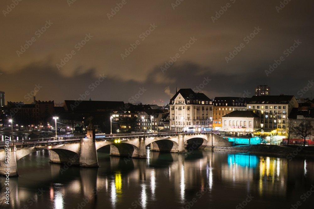 Night view of Basel historical bridge in a smoky Winter night, the Rhine river flows slowly highlighting colorful light reflections on the water surface