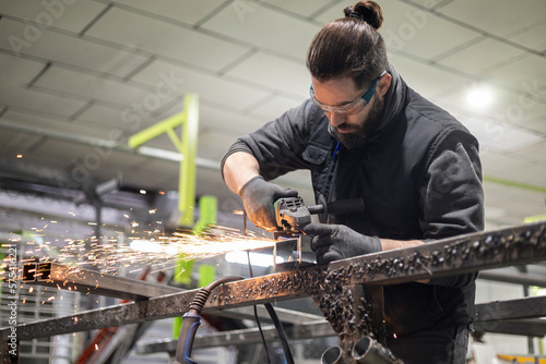 Fotografie Wide shot of a man in protective clothing and glasses using a radial type grinder to cut a metal profile in the workshop