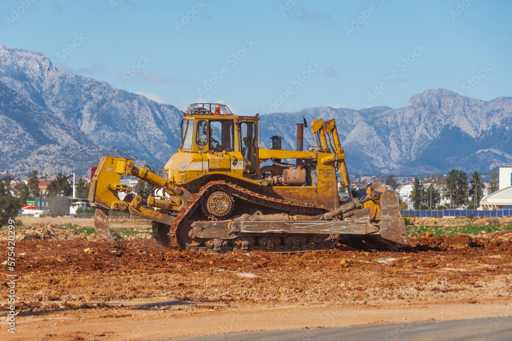 Bulldozer levels the ground in the urban area of a wasteland against the backdrop of mountains.