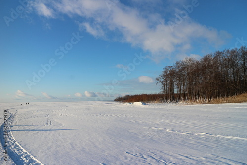 winter frozen lake, shore overgrown with forest, on the snow a trail of sleighs, silhouettes of people in the distance