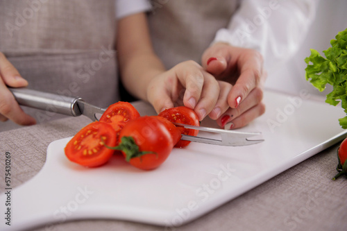 preparing healthy vegetable salad on cutting board in restaurant kitchen. Concept new lean menu close-up of the hands of the child and mother cut cherry tomatoes on a white porcelain board