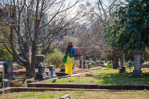 an African American woman wearing a yellow dress, a colorful orange head scarf, a denim jacket with long sisterlocks with lush green trees, headstones and graves at the Oakland Cemetery in Atlanta photo