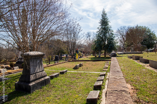 an African American woman wearing a yellow dress, a colorful orange head scarf, a denim jacket with long sisterlocks with lush green trees, headstones and graves at the Oakland Cemetery in Atlanta photo