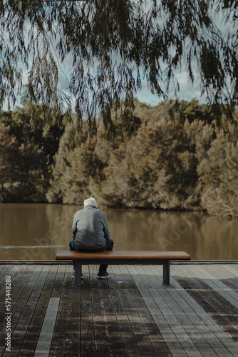 Photography Old man sitting on a bench looking over a pond in a park.