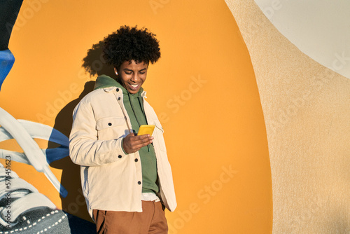 Happy smiling cool gen z young African American ethnic stylish hipster guy model standing at yellow city urban wall using cell phone mobile device, looking at smartphone, holding cellphone. photo